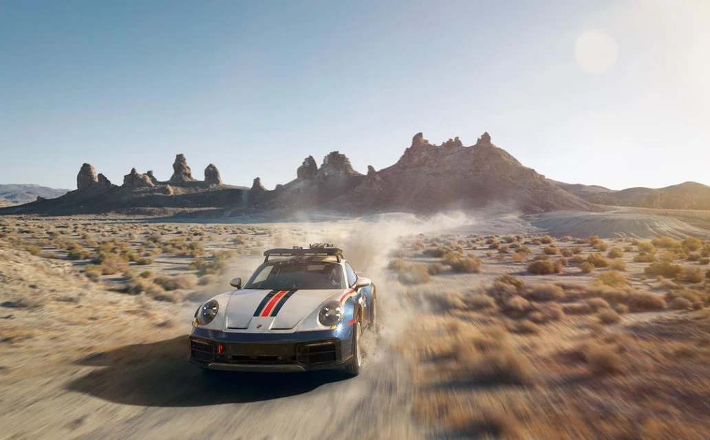 A Porsche 911 Dakar speeds across a desert landscape, kicking up dust with rocky mountains and a bright blue sky in the background.