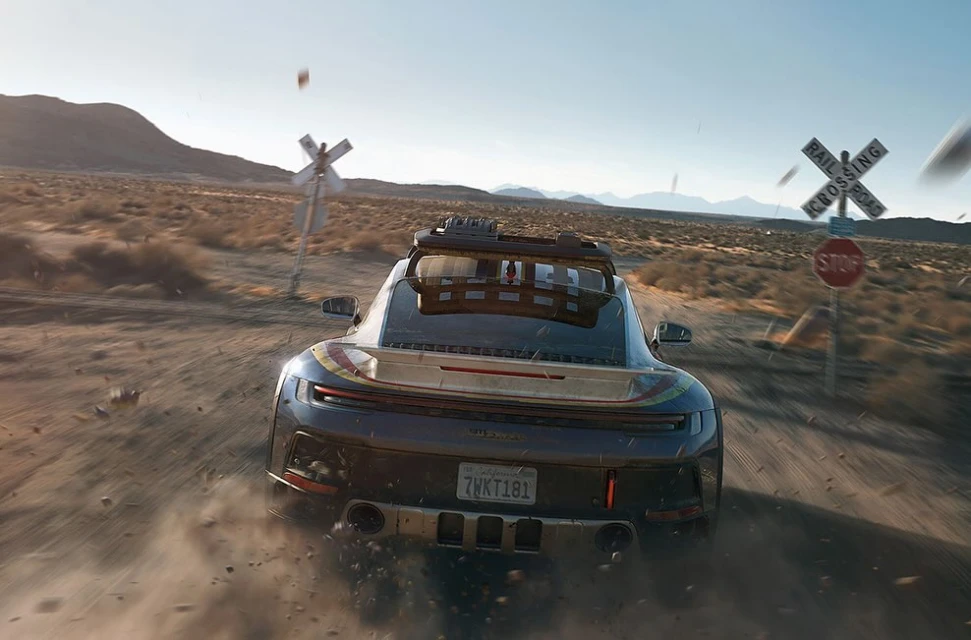 Black car races through a desert landscape near a railroad crossing sign, captured in a dynamic rear-angle shot under bright daylight.