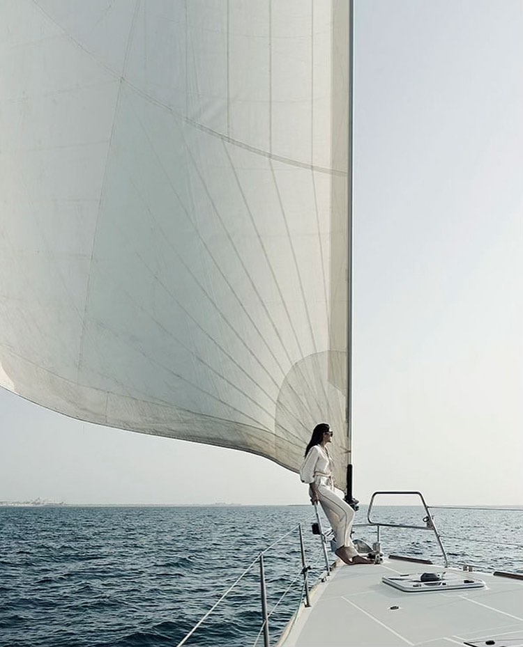 Woman in white clothing on a sailboat with a large sail in the ocean against a clear sky, enjoying a peaceful day sailing.