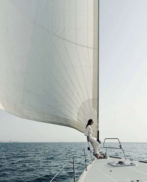 Woman in white clothing on a sailboat with a large sail in the ocean against a clear sky, enjoying a peaceful day sailing.