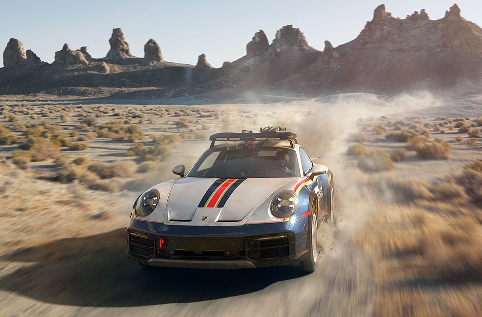 Porsche rally car driving on a dirt road in the desert landscape, with mountains and sparse vegetation in the background.