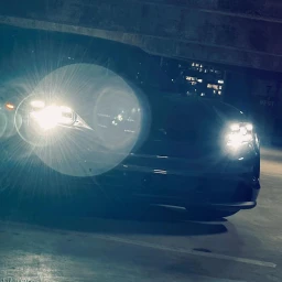 Dark luxury car parked in an underground structure with bright headlights casting strong lens flare.