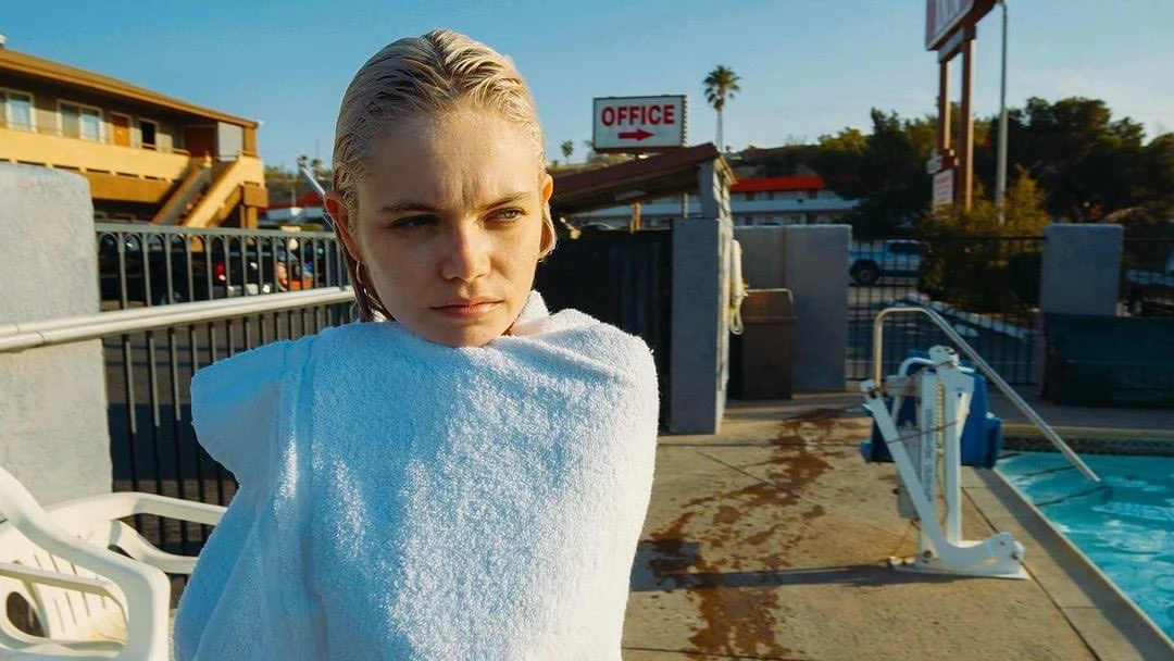 Woman wrapped in a towel sits outdoors in sunlight near a swimming pool in front of a motel with an 'office' sign in the background.