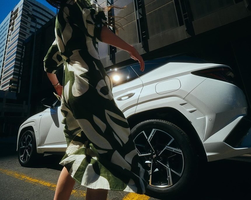 Modern white SUV with geometric design stands out with a woman in patterned dress on an urban street with skyscraper background.