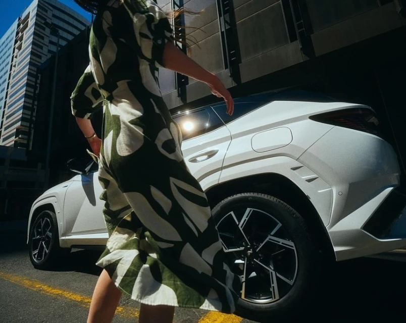 Modern white SUV with geometric design stands out with a woman in patterned dress on an urban street with skyscraper background.