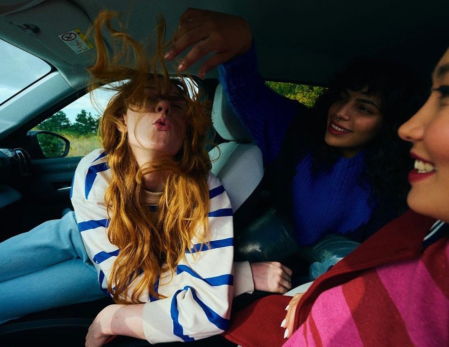 Three women enjoy a car ride, with one playfully posing with her red hair in front of her face, creating a fun, carefree scene. 