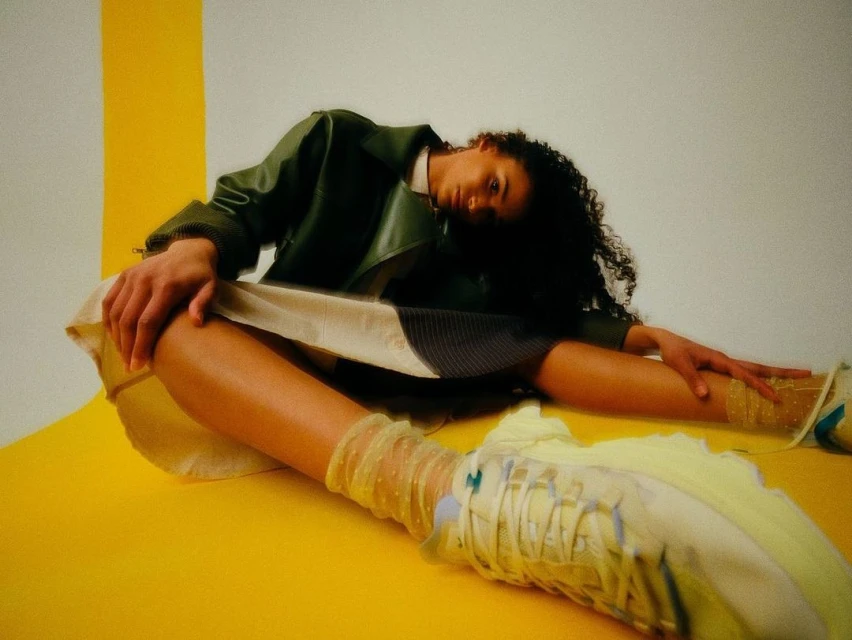 An adult female model in a jacket and skirt poses in a yellow and white photo studio with visible legs, patterned socks, and sneakers.