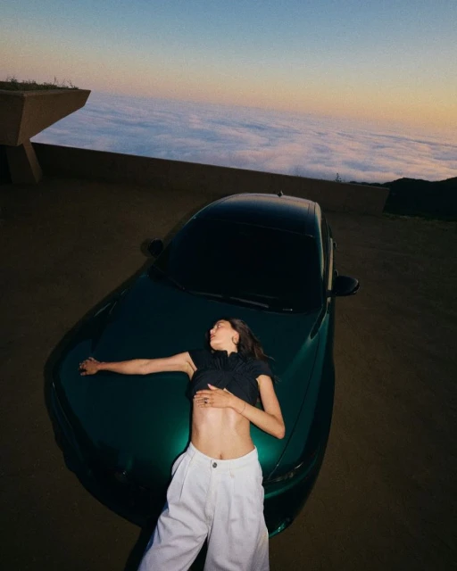 A young woman lies on the hood of a dark green car at dusk, with a sky filled with clouds behind her, captured in a wide shot.
