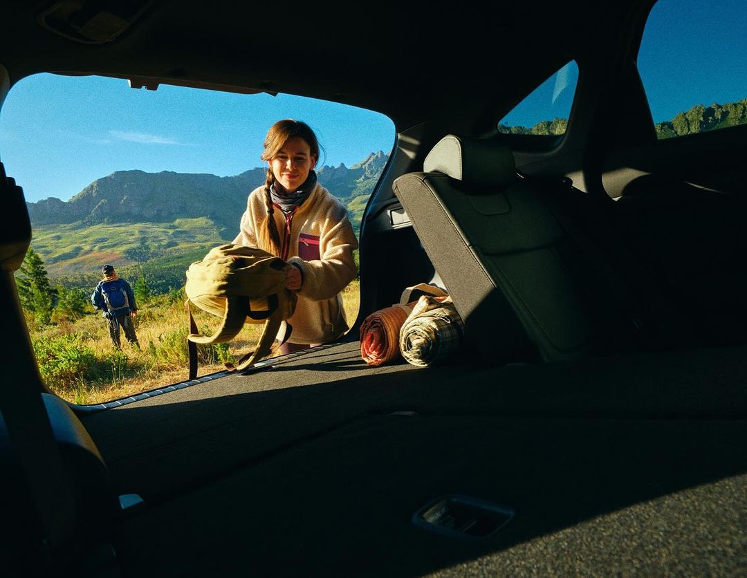 Woman packing backpack into a car trunk with a mountain landscape view. Hiker on a trail in the background. Outdoor adventure.