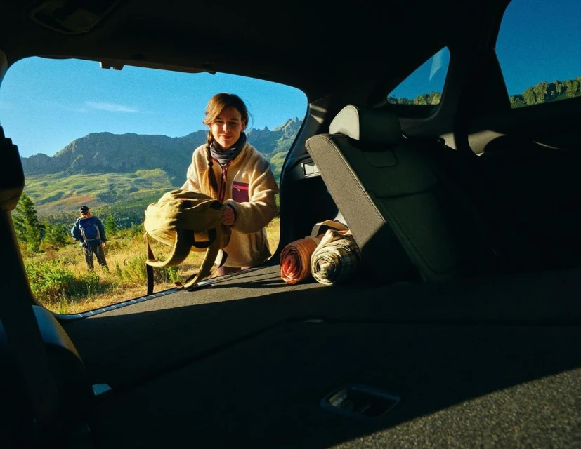 Woman packing backpack into a car trunk with a mountain landscape view. Hiker on a trail in the background. Outdoor adventure.