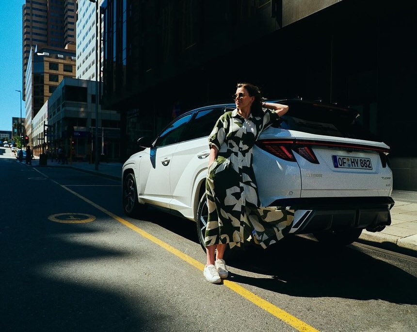 A white SUV is parked on a city street with a woman leaning against the back. Buildings rise in the background on a sunny day.