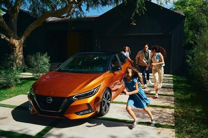 Copper Nissan sedan with a family running cheerfully behind it near their home on a sunny day, capturing a lifestyle moment.