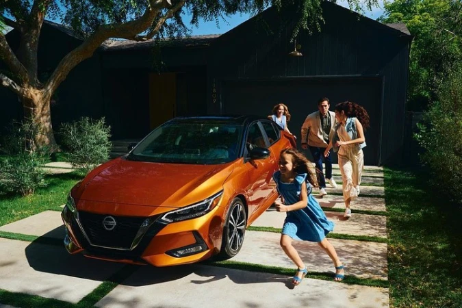 Copper Nissan sedan with a family running cheerfully behind it near their home on a sunny day, capturing a lifestyle moment.