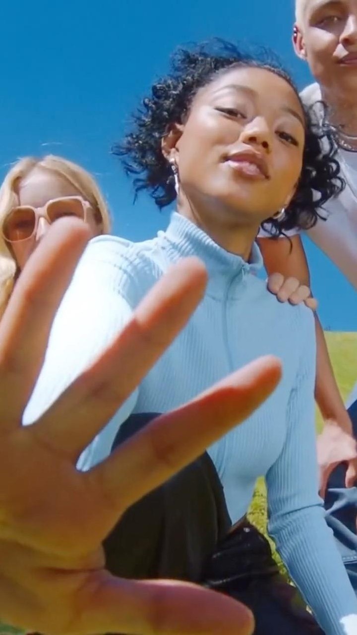 Eye-level shot of a group of three people under blue sky, with one African-descent female in the foreground and green grass in the background.