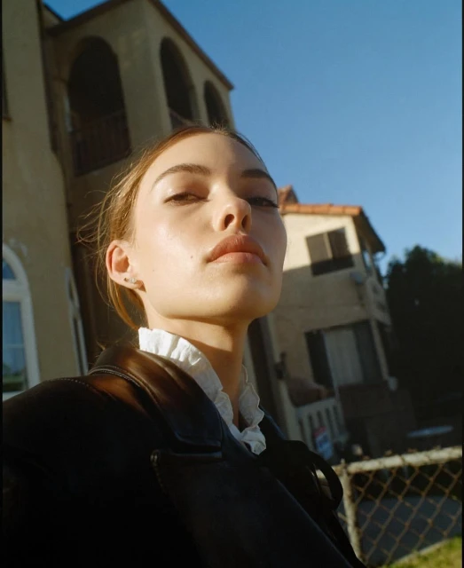 Portrait of a young adult female with fair skin, styled hair, and a black jacket, posing outdoors with architecture in the background.