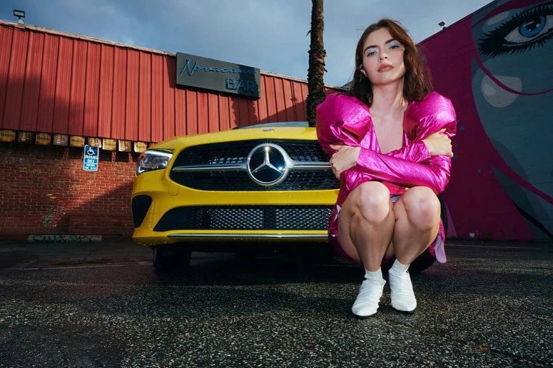 Yellow Mercedes-Benz with an adult female in a pink outfit posing in front of it and a mural on a sunny, paved urban street.
