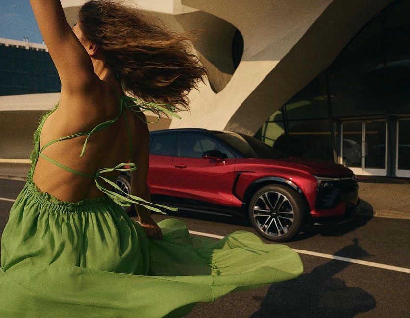 A woman in a flowing green dress stands near a red SUV with a modern building in the background, on a sunny day.