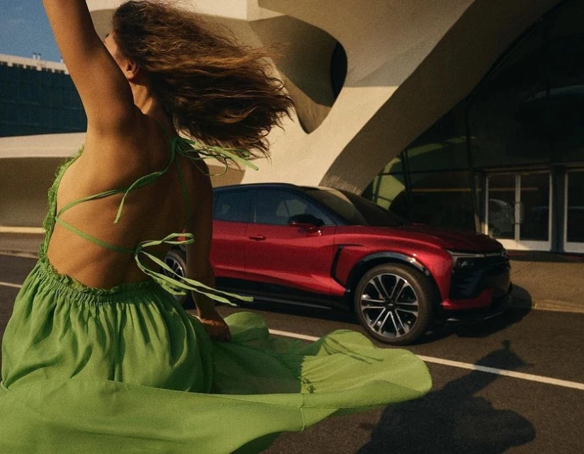 A woman in a flowing green dress stands near a red SUV with a modern building in the background, on a sunny day.