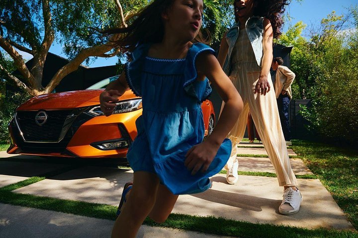 Girl in blue dress runs on a pathway with an orange car, woman in striped jumpsuit, and green trees in the background.