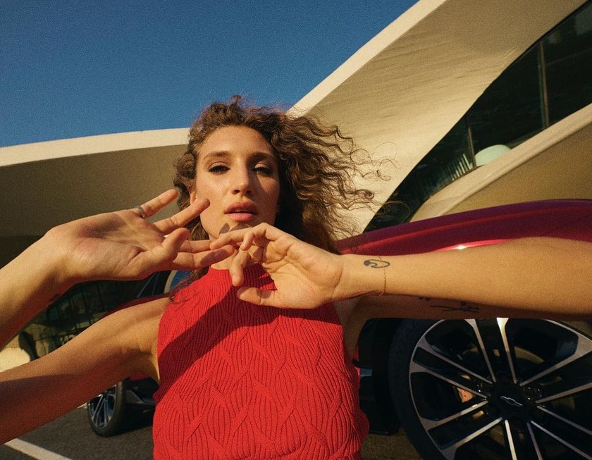 Woman poses playfully outdoors by a red Chevrolet car and modern architecture on a sunny day, with curly hair and a sleeveless red top.