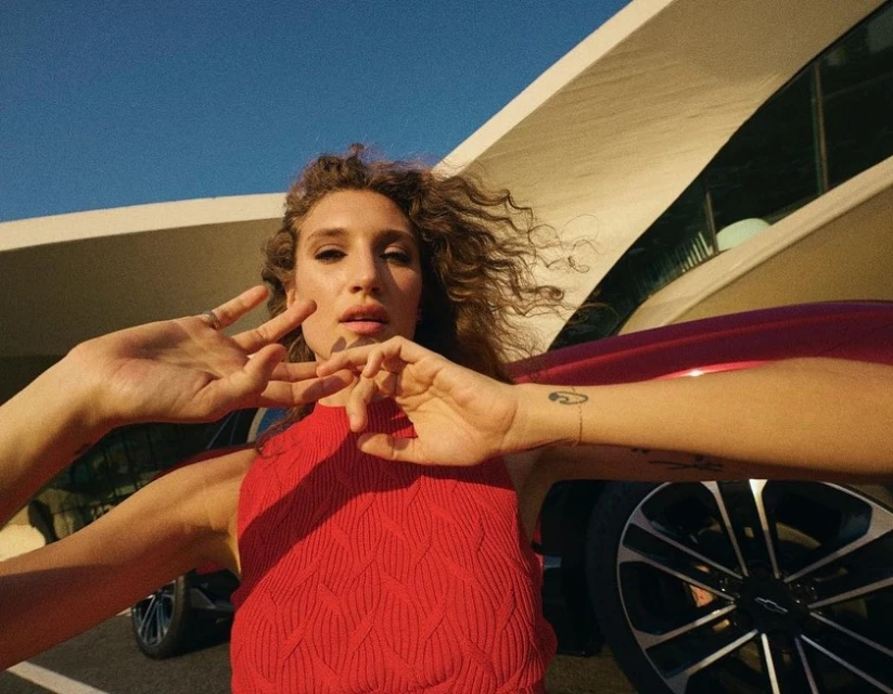 Woman poses playfully outdoors by a red Chevrolet car and modern architecture on a sunny day, with curly hair and a sleeveless red top.