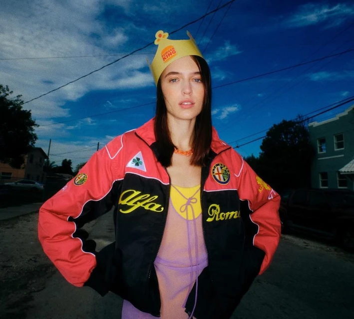Young woman poses in a red and black racing jacket and Burger King crown on a city street with a car and green buildings in background.