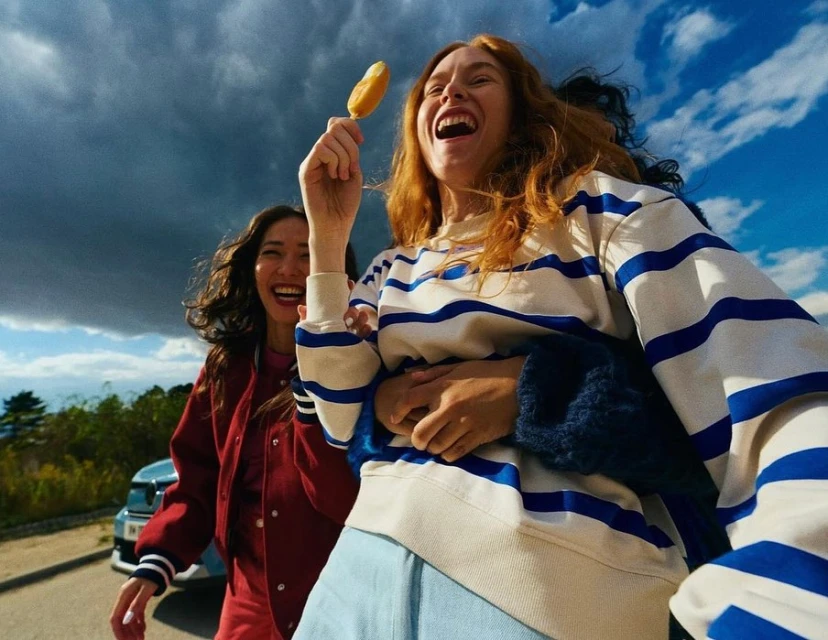 Two women smiling outdoors, one holding a corn dog. The woman in foreground wears a striped top and light jeans against a blue sky.