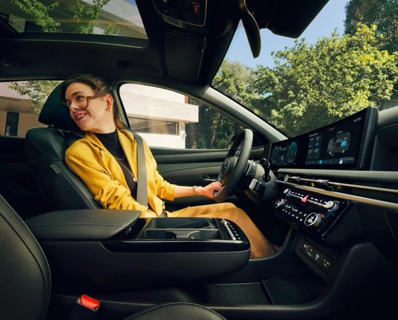 Woman driving a modern car with a large dashboard display, sunroof, and black leather interior; the car is surrounded by trees and buildings.
