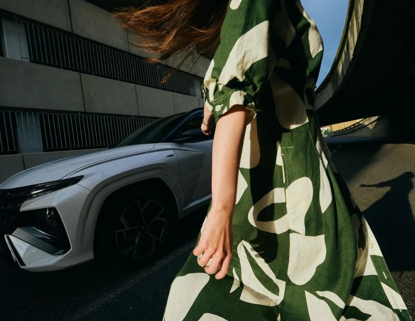 Woman in a green and white dress walks past a white Hyundai Tucson SUV in an urban setting with an overpass and buildings.