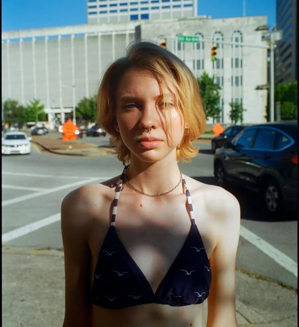 Outdoor portrait of a young adult woman in a swimsuit top, city street background with buildings and cars. Sunny daytime lighting.