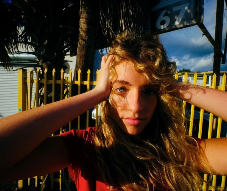 Portrait of a young woman with wavy blonde hair, holding her hands on her head, standing outdoors with a yellow fence in the background.