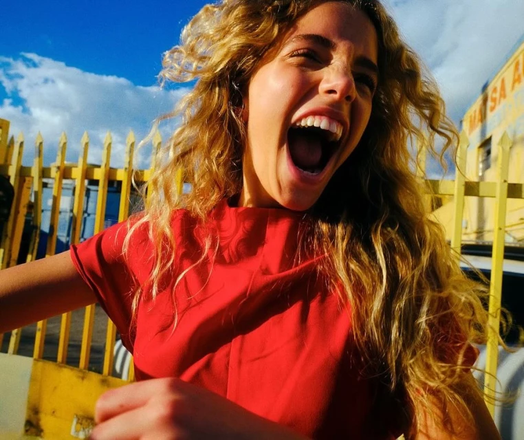 A young woman with long, curly hair wearing a red top stands outdoors in front of a yellow fence, screaming with excitement under a blue sky.