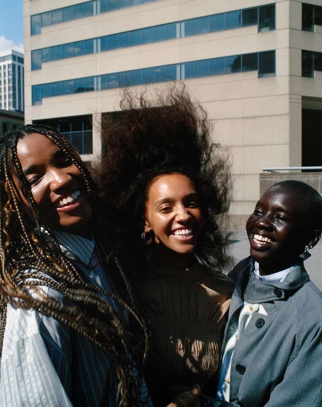 Three smiling women of African descent pose outdoors against a light-colored building, showcasing braided and textured hair in natural daylight.