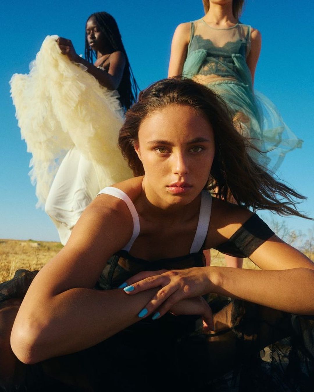 Three women in sheer dresses pose outdoors in a field under a clear sky, capturing a fashion or editorial moment with natural light.