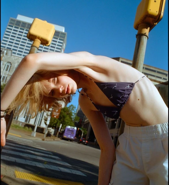 Young woman in a bikini top and pants poses with an extended arm on a city street against a backdrop of buildings and traffic lights.