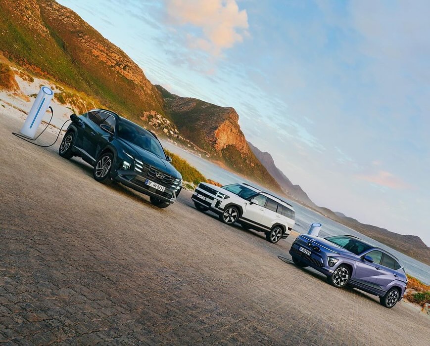 Three electric SUV cars parked on a paved surface with charger stations, mountains, beach and cloudy sky in the background.
