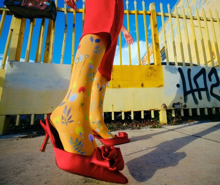 A woman's lower legs and feet are the primary focus, showcasing bright patterned tights and elegant red high heels outdoors.