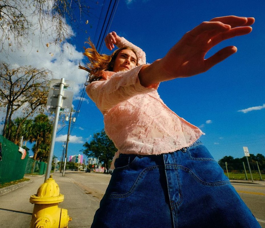 A person in a peach jacket and blue skirt poses on a street next to a yellow fire hydrant on a sunny day with a clear blue sky.