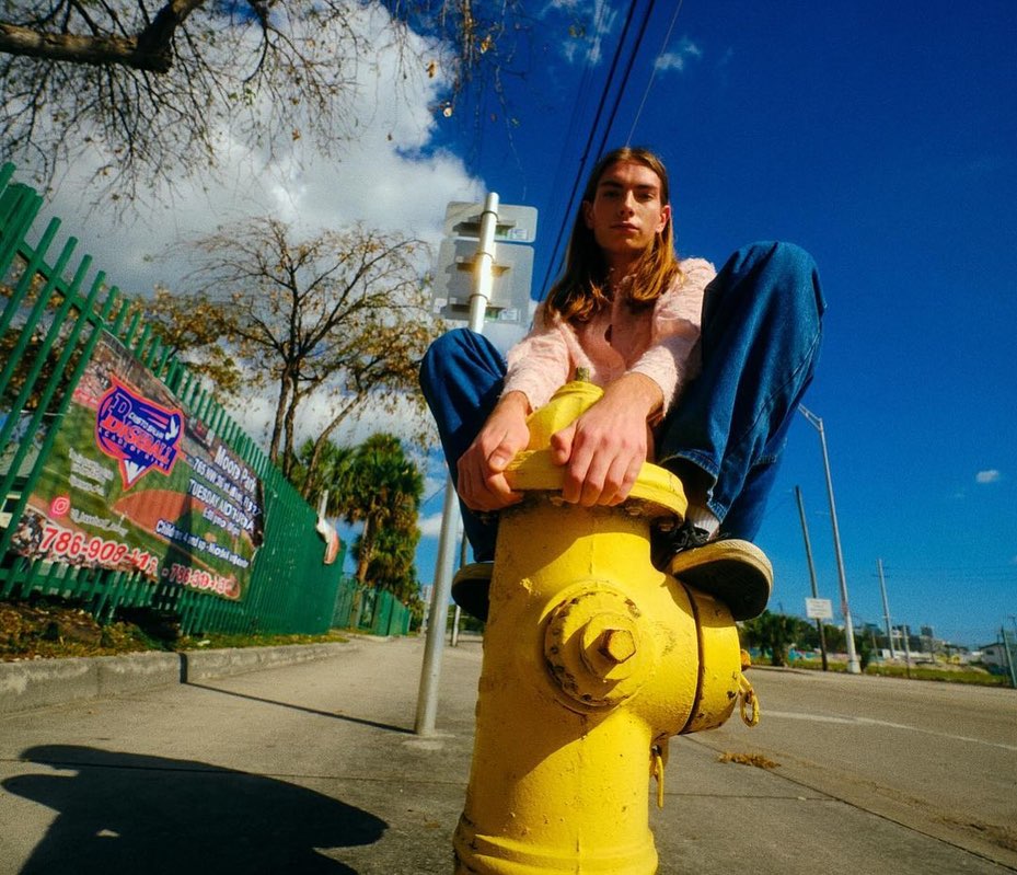 Young adult male sitting on a yellow fire hydrant on a city street under a sunny blue sky wearing jeans and pink sweater.
