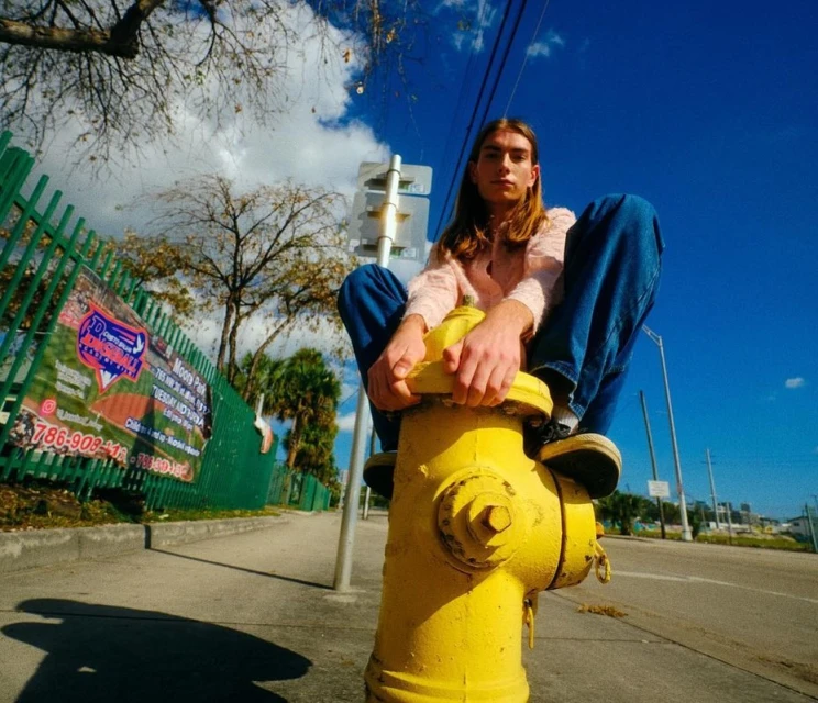 Young adult male sitting on a yellow fire hydrant on a city street under a sunny blue sky wearing jeans and pink sweater.