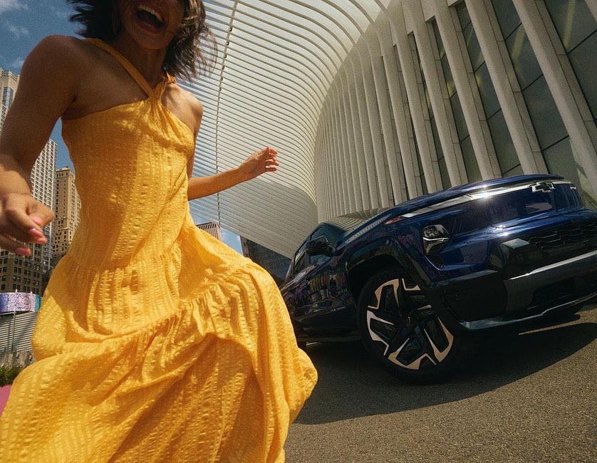 Woman in yellow dress jumping for joy next to blue Chevy truck, cityscape background, capturing a dynamic and vibrant outdoor scene.