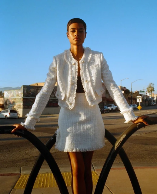Woman in a white tweed skirt suit poses on an urban street with blue sky, capturing fashion and city vibes in bright sunlight.