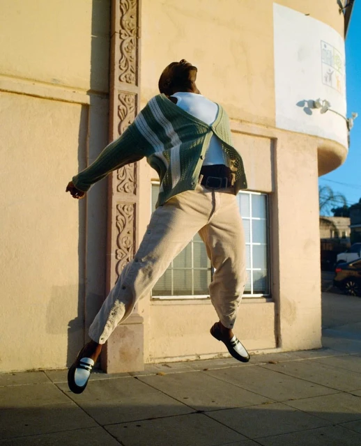 Man in green cardigan and beige pants jumps in front of a building with a window, showcasing a lifestyle fashion image, on city street.