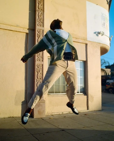 Man in green cardigan and beige pants jumps in front of a building with a window, showcasing a lifestyle fashion image, on city street.