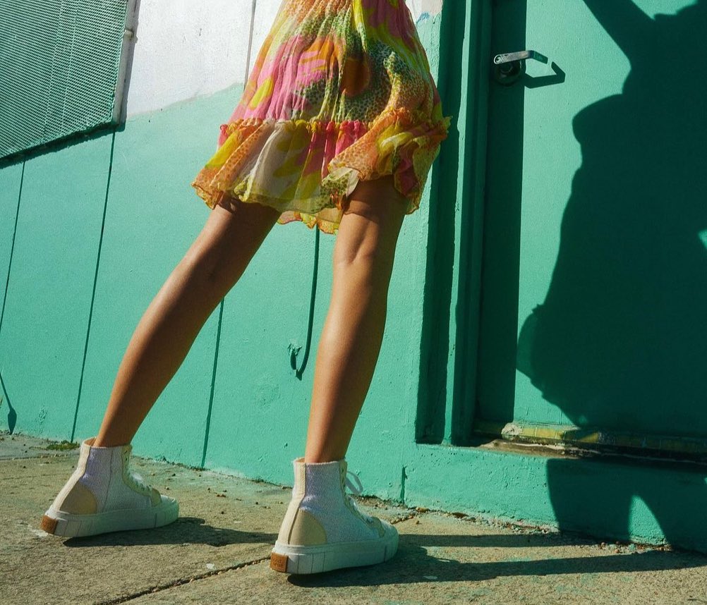 A woman in a floral dress and white high-top sneakers stands in front of a teal building on a sunny day, casting shadows.