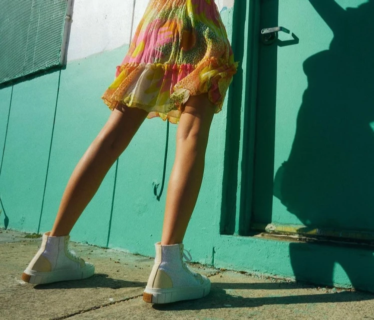 A woman in a floral dress and white high-top sneakers stands in front of a teal building on a sunny day, casting shadows.