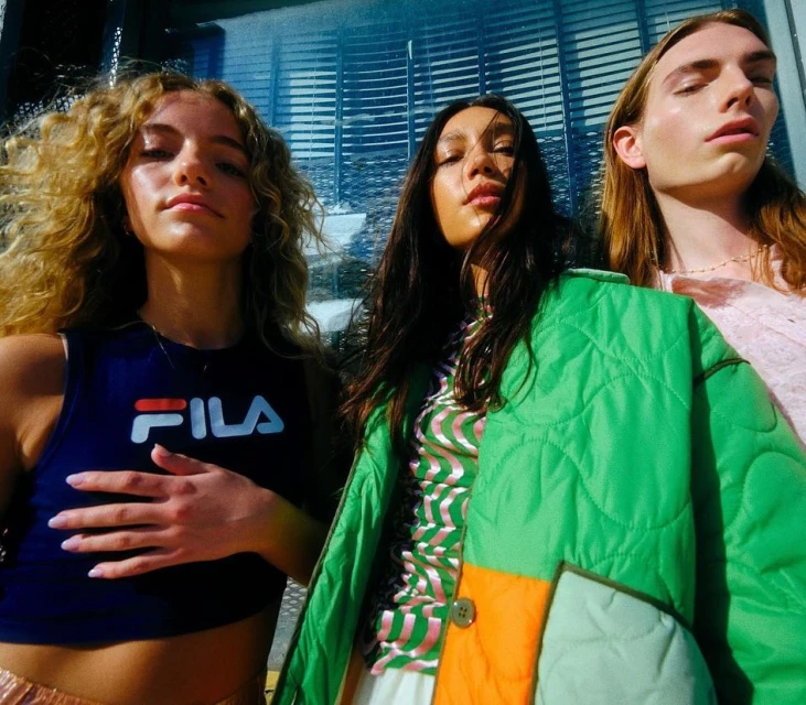 Fashion-forward outdoor portrait of three young adults wearing colorful clothing in sunlight. The models pose against a textured background.