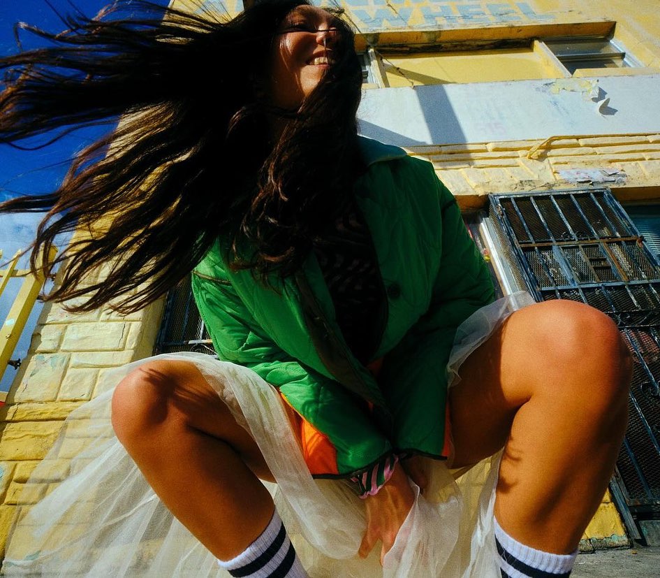 Low-angle shot of an adult woman crouching outdoors with a bright green jacket, white socks, and a translucent white skirt.