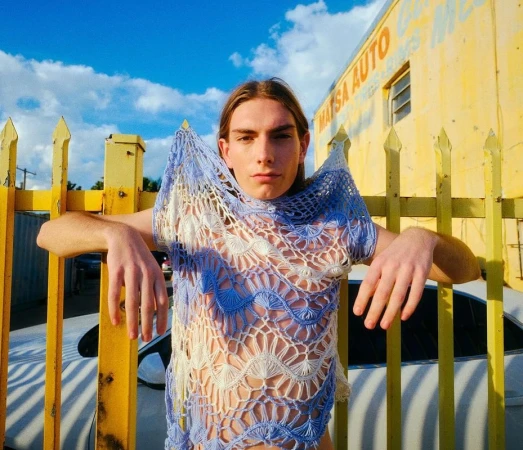 Young adult leans on yellow fence wearing blue crochet top outdoors against blue sky and white car, auto shop in background.