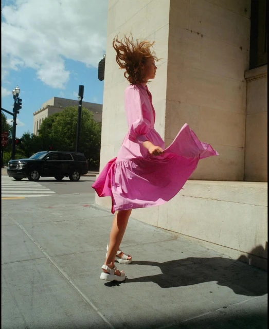 A woman in a pink dress whirls on a city sidewalk next to a concrete wall on a sunny day, with a black SUV in the background.